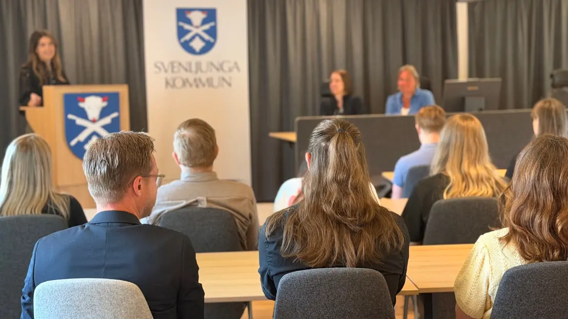 Personer sitter i rader med ryggen vänd mot kameran, framför dem står någon vid en talarstol med Svenljunga kommuns vapen på samt bakom sig, bredvid sitter två personer.