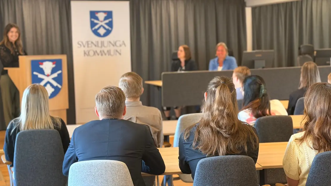 Personer sitter i rader med ryggen vänd mot kameran, framför dem står någon vid en talarstol med Svenljunga kommuns vapen på samt bakom sig, bredvid sitter två personer.