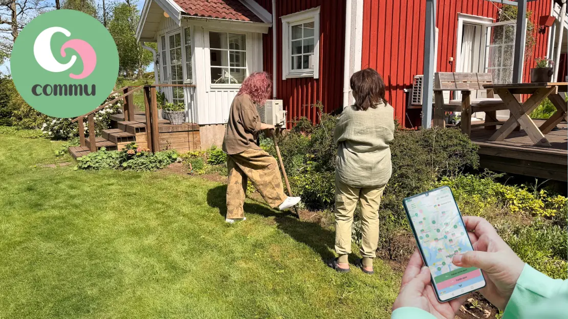 Två personer står vid ett rött trähus, en håller på att gräva med en spade i en blomsterrabatt. I förgrund syns händer som håller en mobil med Commu appen öppen och Commu loggan.