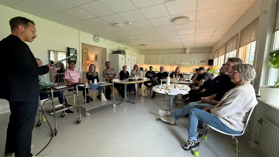 Sebastian står upp och läser ett diplom. Medarbetare sitter i ring framför honom.