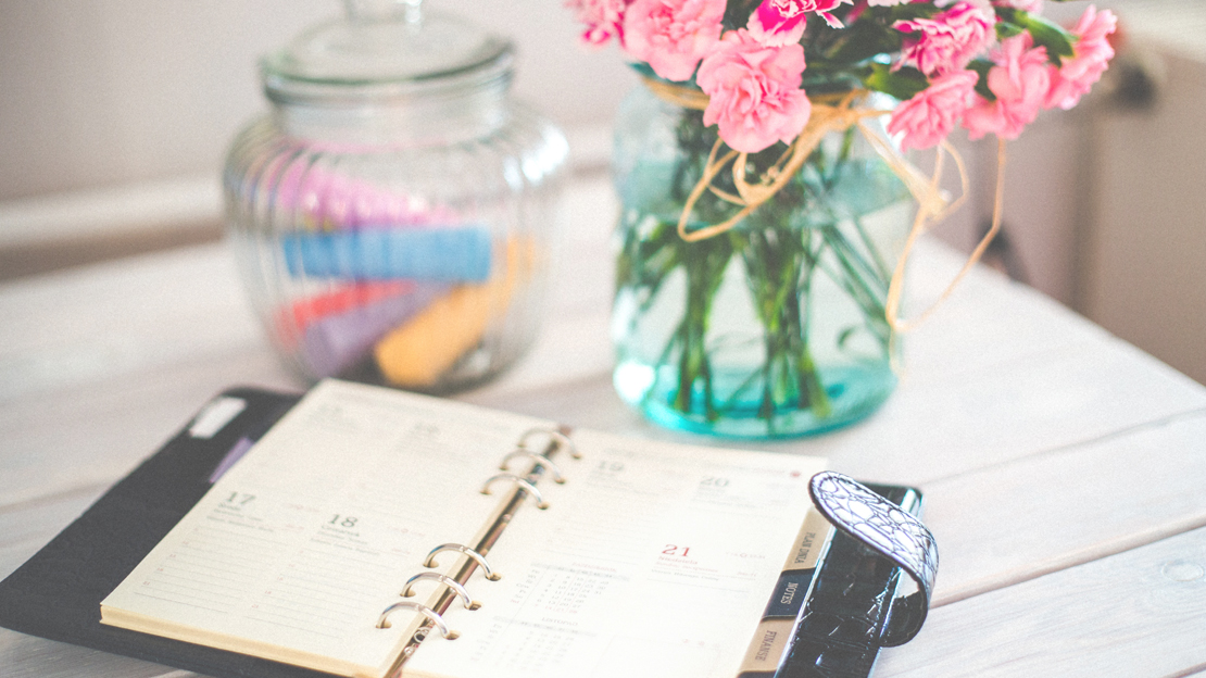 Bild på almanacka som ligger på ett vitt bord tillsammans med en vas med rosa blommor. Länken leder till sida om tider för lov.