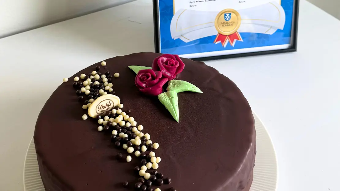 En mörk tårta med rosor på står framför ett diplom med text.