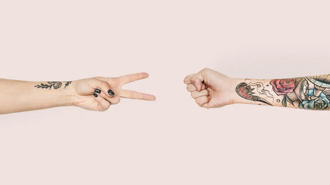 två händer, en gör peacetecken den andra knytnäve, båda armarna är tatuerade