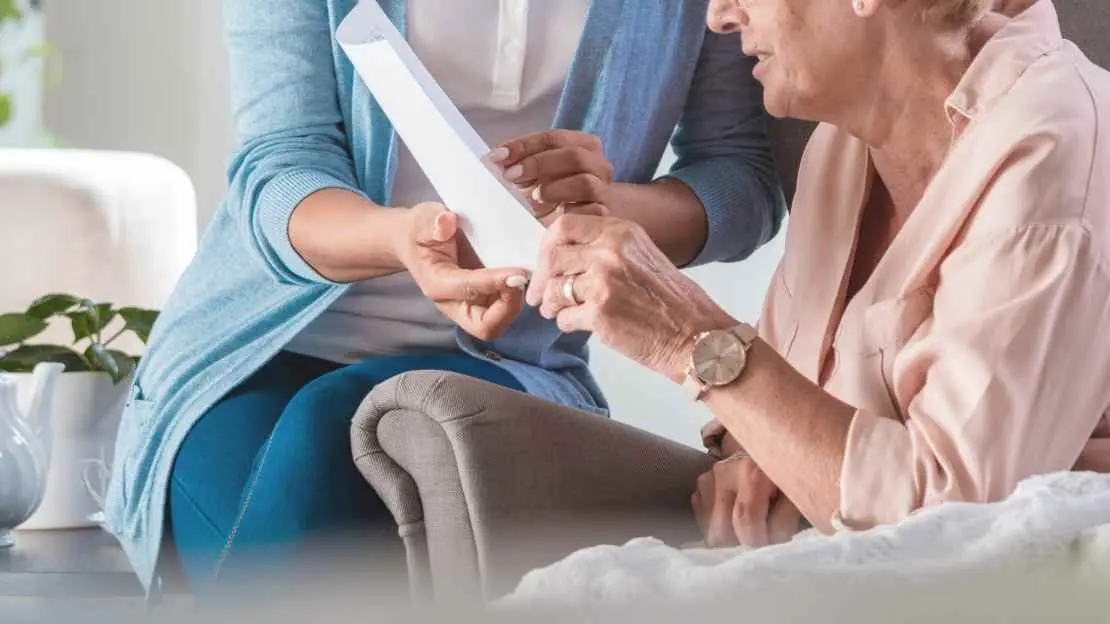 Bild på en äldre person sitter på fåtölj och läser i papper, yngre hjälper att hålla pappret. Länk till vår jobbannons för korttidsvikariat.