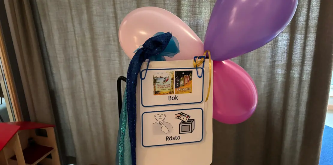 En skylt med röstningscymbol och bild på båda böckerna som bokvalet står emellan. Bakom skylten sitter färgglada ballonger.