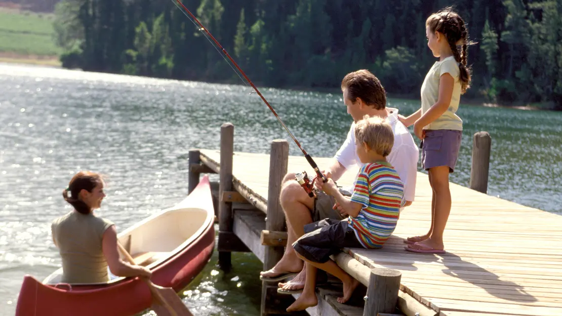 Pappa med två barn är på en badbrygga och fiskar med spö. Jämte bryggan sitter en kvinna i en kajak. 