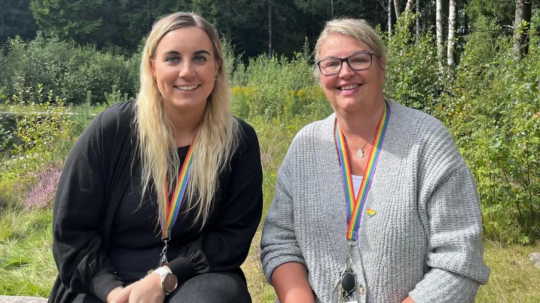 Två kvinnor sitter utomhus och ler mot kameran, båda har nyckelband i pridefärgerna runt halsen.