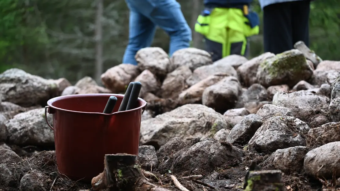 I förgrunden står en hink med två redskap. I bakgrunden finns ett gravröse och man ser nederdelen på tre personer som står bakom röset.