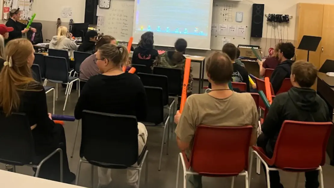 En klassrum med flera personer som sitter på stolar. De tittar på en projektor. De håller Boomwhackers i handen.