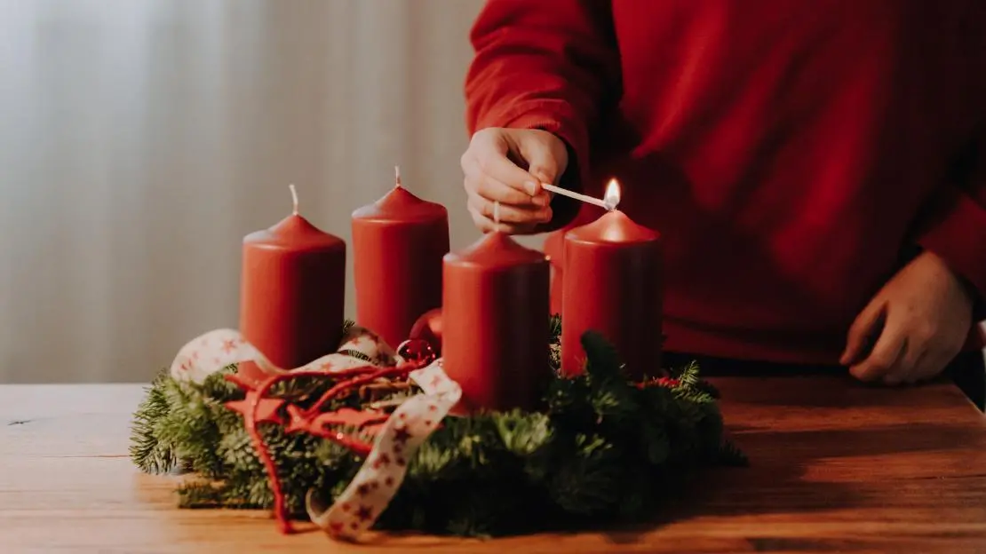 Person i röd tröja tänder ett ljus på en adventskrans med en tändsticka.