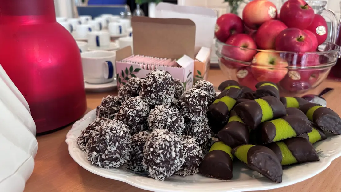 En tallrik med chokladbollar och kakor täckta med choklad och marsipan. En röd kaffekanna och en skål med röda äpplen syns också.