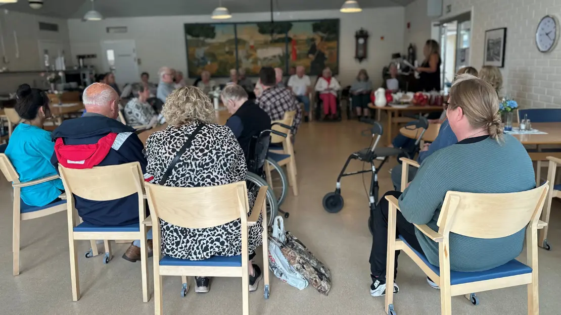 Flera personer sitter på stolar i ett rum. En rullator står i bakgrunden, och en väggdekor visar ett landskap.