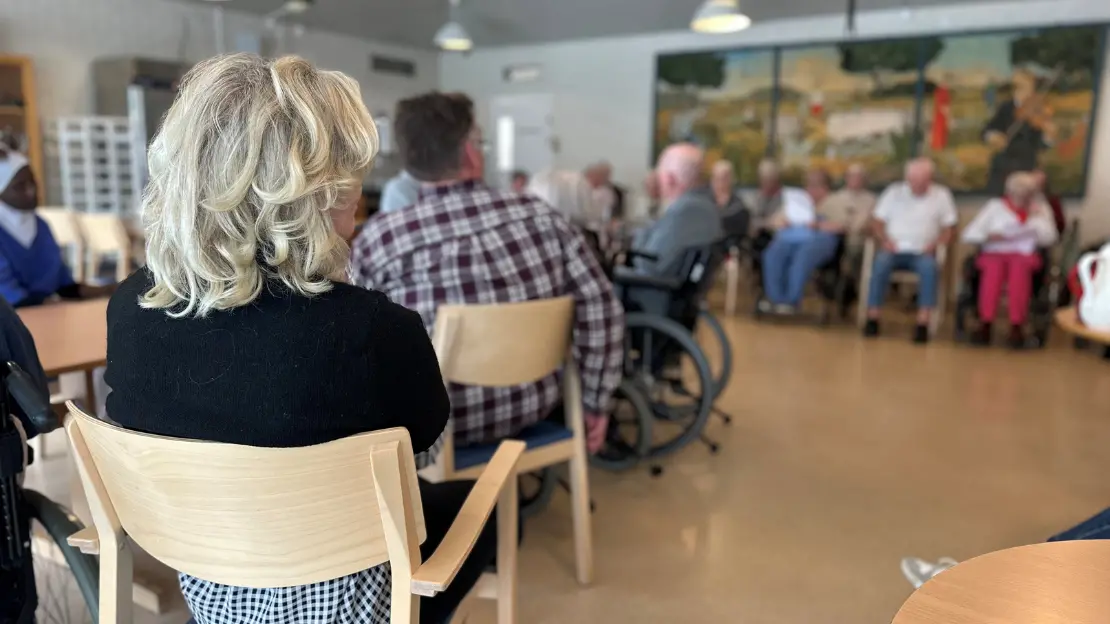 En grupp äldre personer sitter på stolar och rullstolar i ett rum. En tavla med ett landskap hänger på väggen bakom.