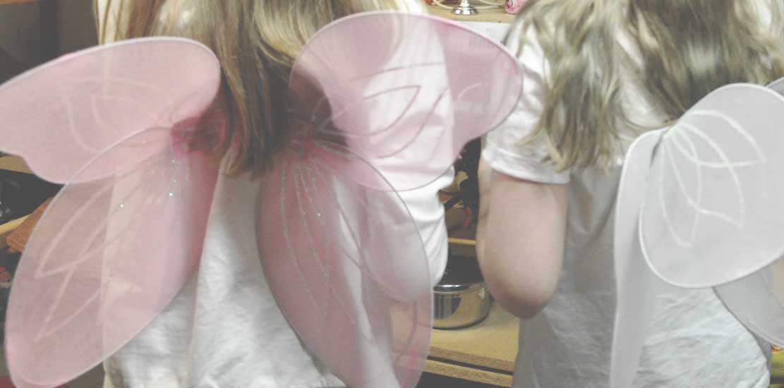 Två barn som står med ryggarna vända mot kameran och på ryggen har dom rosa och vita vingar