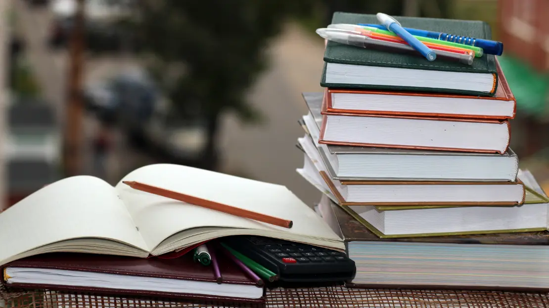 på en bänk ligger en hög med böcker en öppen skrivbok tillsammans med några pennor och en miniräknare.