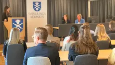 Personer sitter i rader med ryggen vänd mot kameran, framför dem står någon vid en talarstol med Svenljunga kommuns vapen på samt bakom sig, bredvid sitter två personer.