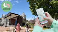 Två personer går över bron i lilla parken vid torget. I förgrund syns händer som håller en mobil med Commu appen öppen och Commu loggan.