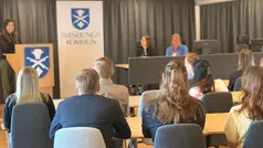 Personer sitter i rader med ryggen vänd mot kameran, framför dem står någon vid en talarstol med Svenljunga kommuns vapen på samt bakom sig, bredvid sitter två personer.