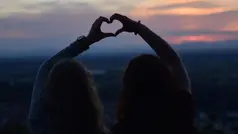 Två personer bildar ett hjärta med sina händer framför en solnedgång.
