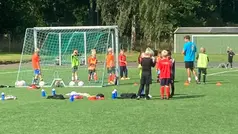 Flera barn står vid ett fotbollsmål och har träning med en ungdom som ledare.