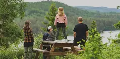 En familj står vid och på ett picknickbord med utsikt över en sjö.