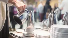 Person häller upp vätska ur en kanna in i en kopp vid ett bord med många tallrikar och kannor.