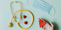 på ljusblå bakgrund ligger ett gult stetoskop två små röda hjärtan och ett blått munskydd, från nedre högra hörnet strcäker sig en hand in i bilden som håller i ett rött pappershjärta