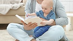 Person sitter på golvet med baby i famnen och håller en bok som bebisen bläddrar i.