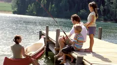 Pappa med två barn är på en badbrygga och fiskar med spö. Jämte bryggan sitter en kvinna i en kajak. 