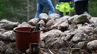 I förgrunden står en hink med två redskap. I bakgrunden finns ett gravröse och man ser nederdelen på tre personer som står bakom röset.