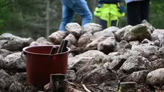 I förgrunden står en hink med två redskap. I bakgrunden finns ett gravröse och man ser nederdelen på tre personer som står bakom röset.