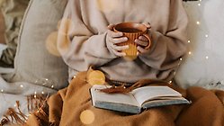 Person i tjocktröja sitter med kopp i handen och läser en bok med ett höstlöv på. 