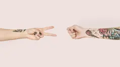 två händer, en gör peacetecken den andra knytnäve, båda armarna är tatuerade