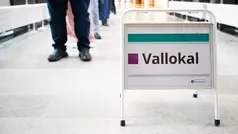 En skylt med texten "Vallokal" i ett stativ. Personer syns gående i bakgrunden.