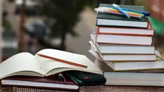 på en bänk ligger en hög med böcker en öppen skrivbok tillsammans med några pennor och en miniräknare.