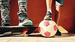 röd vägg utomhus framför syns fötter på två barn i träningsskor, ena barnet har en fot på en skateboard, andra barnet har en fot på en fotboll rosa och vita rutot.